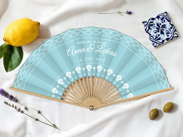 Personalisierter Hochzeitsfächer „Am Meer“ mit den Namen Anna & Lukas, Wellen am Rand und Muschelbogen unter dem Schriftzug, Stoff in Himmelblau mit Druck in Weiß, auf einem Hochzeitstisch