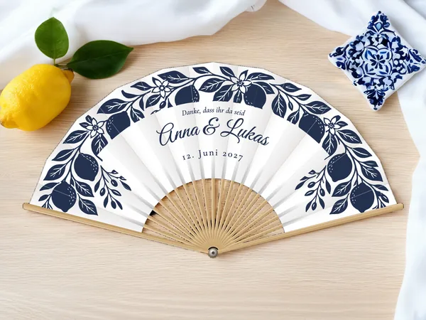 Personalisierter Hochzeitsfächer „Amalfi-Zitronen“ mit den Namen Anna & Lukas, Zitronenzweige und Blüten um den Schriftzug, Stoff in Weiß mit Druck in Marineblau, auf einem Hochzeitstisch