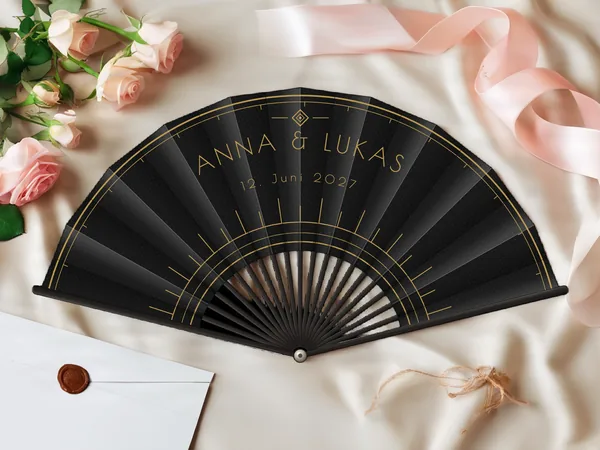 Personalisierter Hochzeitsfächer „Art déco“ mit den Namen Anna & Lukas, feine Bögen und grafische Art-déco-Strahlen um die Mitte, Stoff in Schwarz mit Druck in Gold, auf einem Hochzeitstisch