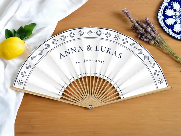 Personalisierter Hochzeitsfächer „Azulejo“ mit den Namen Anna & Lukas, Fries aus rautenförmigen Fliesen entlang des Randes, Stoff in Weiß mit Druck in Marineblau, auf einem Hochzeitstisch