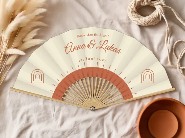 Personalisierter Hochzeitsfächer „Boho-Sonne“ mit den Namen Anna & Lukas, strahlende halbe Sonne unter dem Schriftzug und kleine Bögen an den Seiten, Stoff in Elfenbein mit Druck in Terrakotta, auf einem Hochzeitstisch