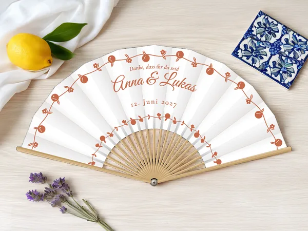 Personalisierter Hochzeitsfächer „Dolce Vita“ mit den Namen Anna & Lukas, Girlanden aus kleinen Zitrusfrüchten und Blüten in Bögen um den Schriftzug, Stoff in Weiß mit Druck in Terrakotta, auf einem Hochzeitstisch