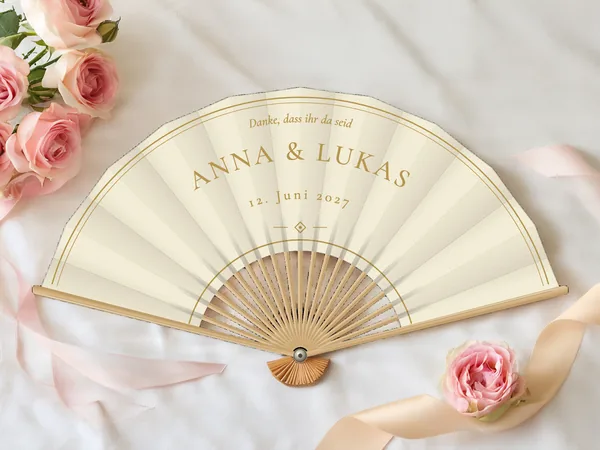 Personalisierter Hochzeitsfächer „Elfenbein und Gold“ mit den Namen Anna & Lukas, doppelte feine Randlinie und kleine Raute unter dem Schriftzug, Stoff in Elfenbein mit Druck in Gold, auf einem Hochzeitstisch
