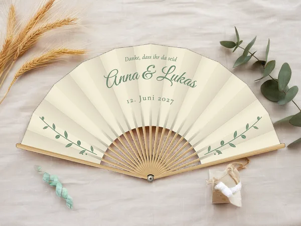 Personalisierter Hochzeitsfächer „Eukalyptus“ mit den Namen Anna & Lukas, feine belaubte Zweige zu beiden Seiten unter dem Schriftzug, Stoff in Elfenbein mit Druck in Salbeigrün, auf einem Hochzeitstisch