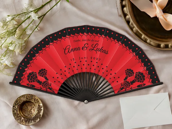Personalisierter Hochzeitsfächer „Flamenco“ mit den Namen Anna & Lukas, gepunkteter Spitzenvolant am Rand und Nelken in den Ecken, Stoff in Rot mit Druck in Schwarz, auf einem Hochzeitstisch