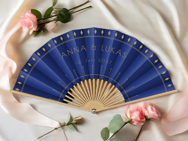 Personalisierter Hochzeitsfächer „Gatsby“ mit den Namen Anna & Lukas, Rauten entlang des Randes und Art-déco-Strahlen unter dem Schriftzug, Stoff in Marineblau mit Druck in Gold, auf einem Hochzeitstisch