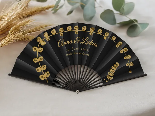 Personalisierter Hochzeitsfächer „Goldener Eukalyptus“ mit den Namen Anna & Lukas, Eukalyptuszweige im Bogen um den Schriftzug, Stoff in Schwarz mit Druck in Gold, auf einem Hochzeitstisch