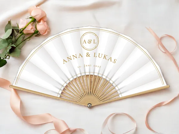 Personalisierter Hochzeitsfächer „Goldenes Monogramm“ mit den Namen Anna & Lukas, Medaillon mit Initialen über dem Schriftzug und doppelte Randlinie, Stoff in Weiß mit Druck in Gold, auf einem Hochzeitstisch