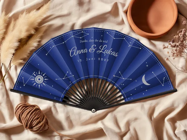 Personalisierter Hochzeitsfächer „Himmelszelt“ mit den Namen Anna & Lukas, Sternbilder, Sonne und Mondsichel rund um den Schriftzug, Stoff in Marineblau mit Druck in Silber, auf einem Hochzeitstisch