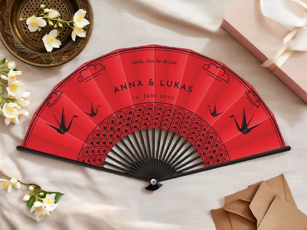 Personalisierter Hochzeitsfächer „Kimono“ mit den Namen Anna & Lukas, Origami-Kraniche, Wolken und Wellenmuster um die Mitte, Stoff in Rot mit Druck in Schwarz, auf einem Hochzeitstisch