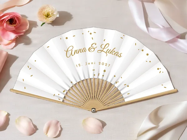 Personalisierter Hochzeitsfächer „Konfetti“ mit den Namen Anna & Lukas, kleines Konfetti rund um den Schriftzug verstreut, Stoff in Weiß mit Druck in Gold, auf einem Hochzeitstisch