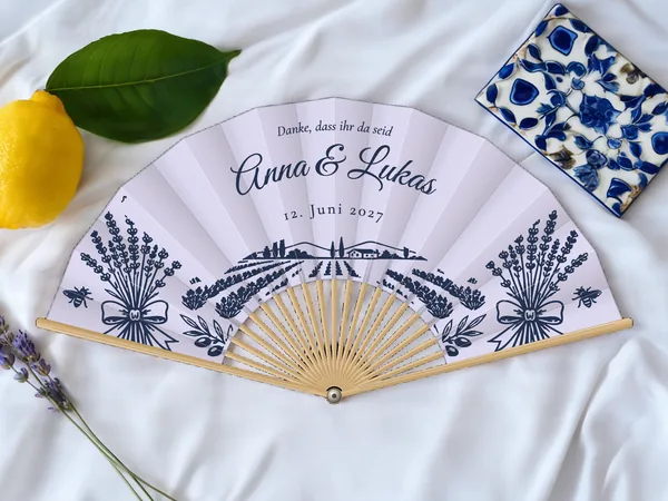 Personalisierter Hochzeitsfächer „Lavendelfeld“ mit den Namen Anna & Lukas, Lavendelsträuße, Bienen und Lavendelfeld unter dem Schriftzug, Stoff in Lavendel mit Druck in Marineblau, auf einem Hochzeitstisch