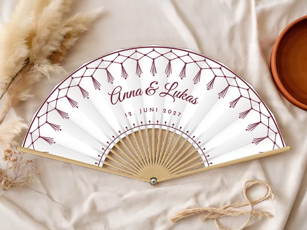 Personalisierter Hochzeitsfächer „Makramee“ mit den Namen Anna & Lukas, Makrameenetz mit Quasten entlang des Randes, Stoff in Weiß mit Druck in Bordeaux, auf einem Hochzeitstisch