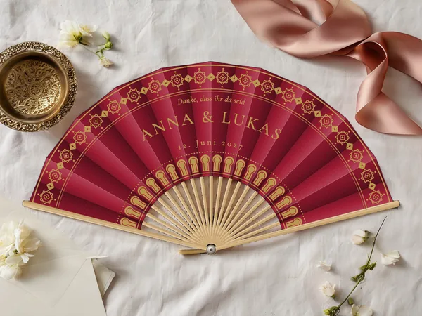 Personalisierter Hochzeitsfächer „Medina“ mit den Namen Anna & Lukas, Fries aus orientalischen Rosetten am Rand und Arkaden um die Mitte, Stoff in Bordeaux mit Druck in Gold, auf einem Hochzeitstisch