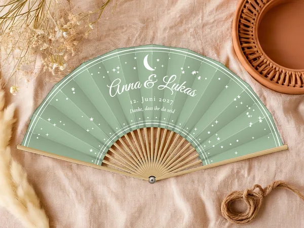 Personalisierter Hochzeitsfächer „Mond und Sterne“ mit den Namen Anna & Lukas, Mondsichel und verstreute Sterne um den Schriftzug, Stoff in Salbeigrün mit Druck in Weiß, auf einem Hochzeitstisch