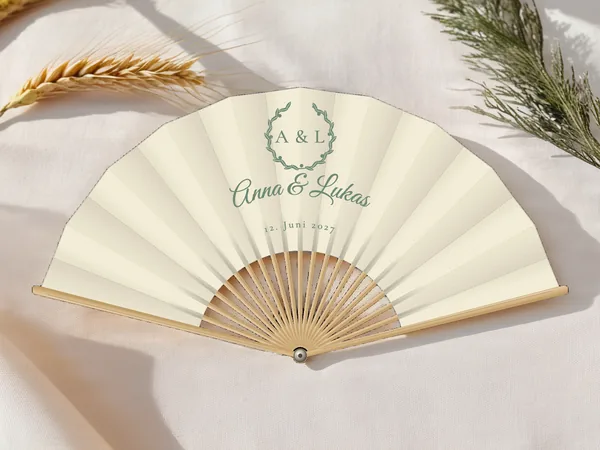 Personalisierter Hochzeitsfächer „Olivenkranz“ mit den Namen Anna & Lukas, Blätterkranz um die Initialen über dem Schriftzug, Stoff in Elfenbein mit Druck in Salbeigrün, auf einem Hochzeitstisch