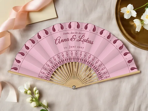 Personalisierter Hochzeitsfächer „Paisley“ mit den Namen Anna & Lukas, Paisleytropfen entlang des Randes und Rosette um die Mitte, Stoff in Puderrosa mit Druck in Bordeaux, auf einem Hochzeitstisch