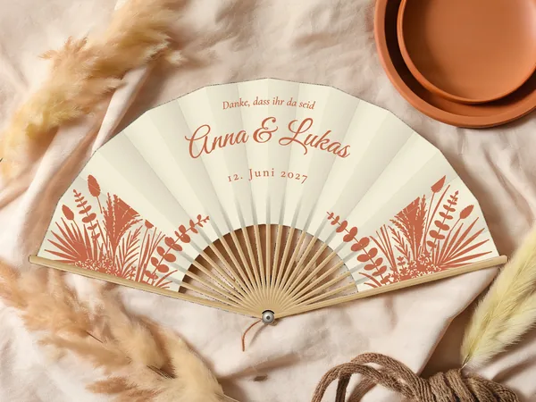 Personalisierter Hochzeitsfächer „Pampasgras“ mit den Namen Anna & Lukas, Pampasgras, Palmwedel und Trockengräser in den unteren Ecken, Stoff in Elfenbein mit Druck in Terrakotta, auf einem Hochzeitstisch