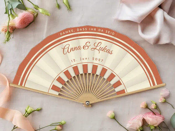 Personalisierter Hochzeitsfächer „Regenbogen“ mit den Namen Anna & Lukas, breite Bogenstreifen am Rand und Zinnen um die Mitte, Stoff in Elfenbein mit Druck in Terrakotta, auf einem Hochzeitstisch