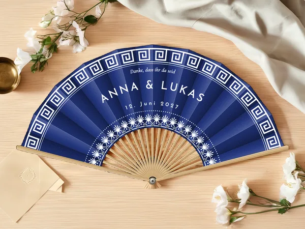 Personalisierter Hochzeitsfächer „Santorini“ mit den Namen Anna & Lukas, griechischer Mäanderfries am Rand und Blumenkranz um die Mitte, Stoff in Marineblau mit Druck in Weiß, auf einem Hochzeitstisch