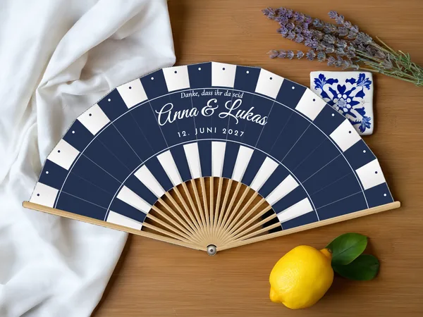 Personalisierter Hochzeitsfächer „Sonnenschirm“ mit den Namen Anna & Lukas, Sonnenschirmstreifen als Bänder am Rand und unten, Stoff in Weiß mit Druck in Marineblau, auf einem Hochzeitstisch