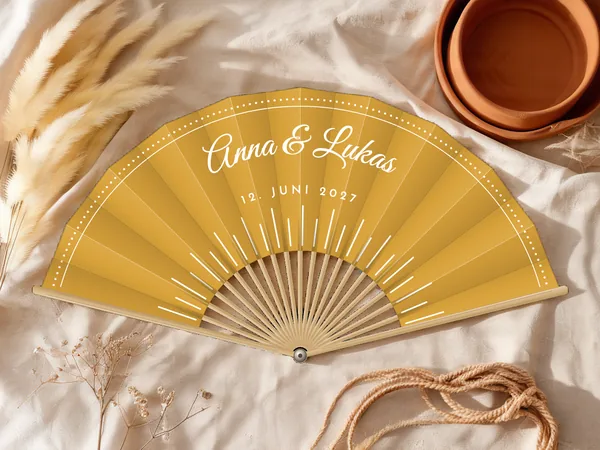 Personalisierter Hochzeitsfächer „Sonnenstrahlen“ mit den Namen Anna & Lukas, feine Strahlen von unten und gepunkteter Fries am Rand, Stoff in Senfgelb mit Druck in Weiß, auf einem Hochzeitstisch