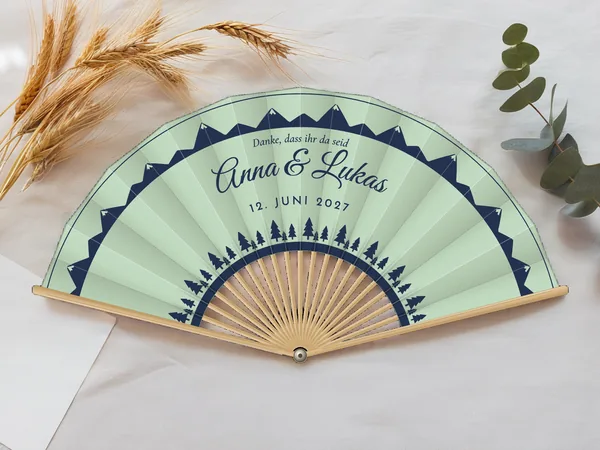 Personalisierter Hochzeitsfächer „Tannen und Berge“ mit den Namen Anna & Lukas, Bergkämme am Rand und eine Reihe Tannen unten, Stoff in Mintgrün mit Druck in Marineblau, auf einem Hochzeitstisch