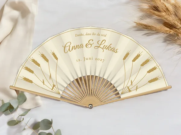 Personalisierter Hochzeitsfächer „Weizenähren“ mit den Namen Anna & Lukas, aufrechte Weizenähren zu beiden Seiten und feine Randlinie, Stoff in Elfenbein mit Druck in Gold, auf einem Hochzeitstisch
