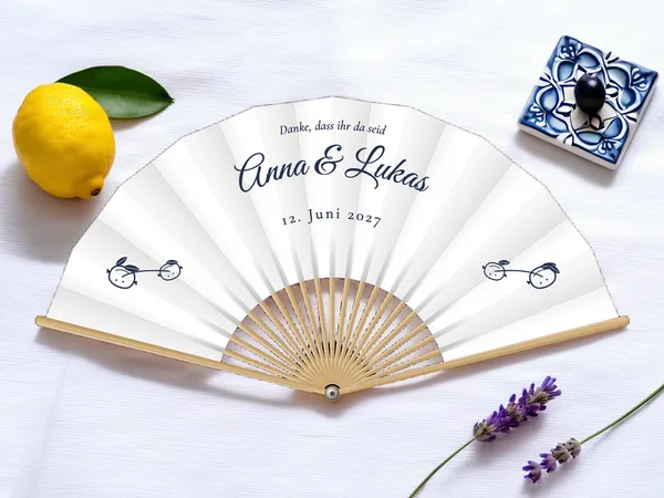 Personalisierter Hochzeitsfächer „Zitronenbaum“ mit den Namen Anna & Lukas, kleiner Zweig mit zwei Zitronen zu beiden Seiten unter dem Schriftzug, Stoff in Weiß mit Druck in Marineblau, auf einem Hochzeitstisch